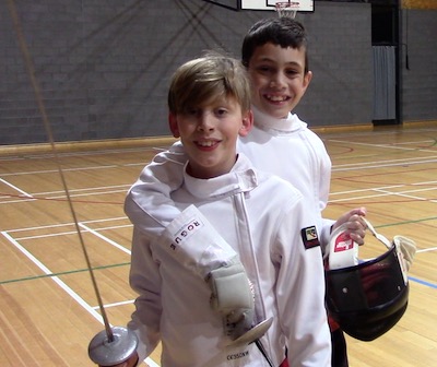 Two boys, aged about 11 and 9, in fencing gear; one has his arm around the other's shoulders