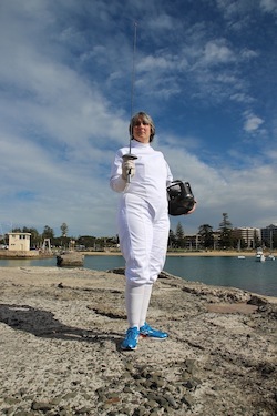 A photo of Coach Laura standing on a rocky beach dressed in fencing whites and holding a sword and mask